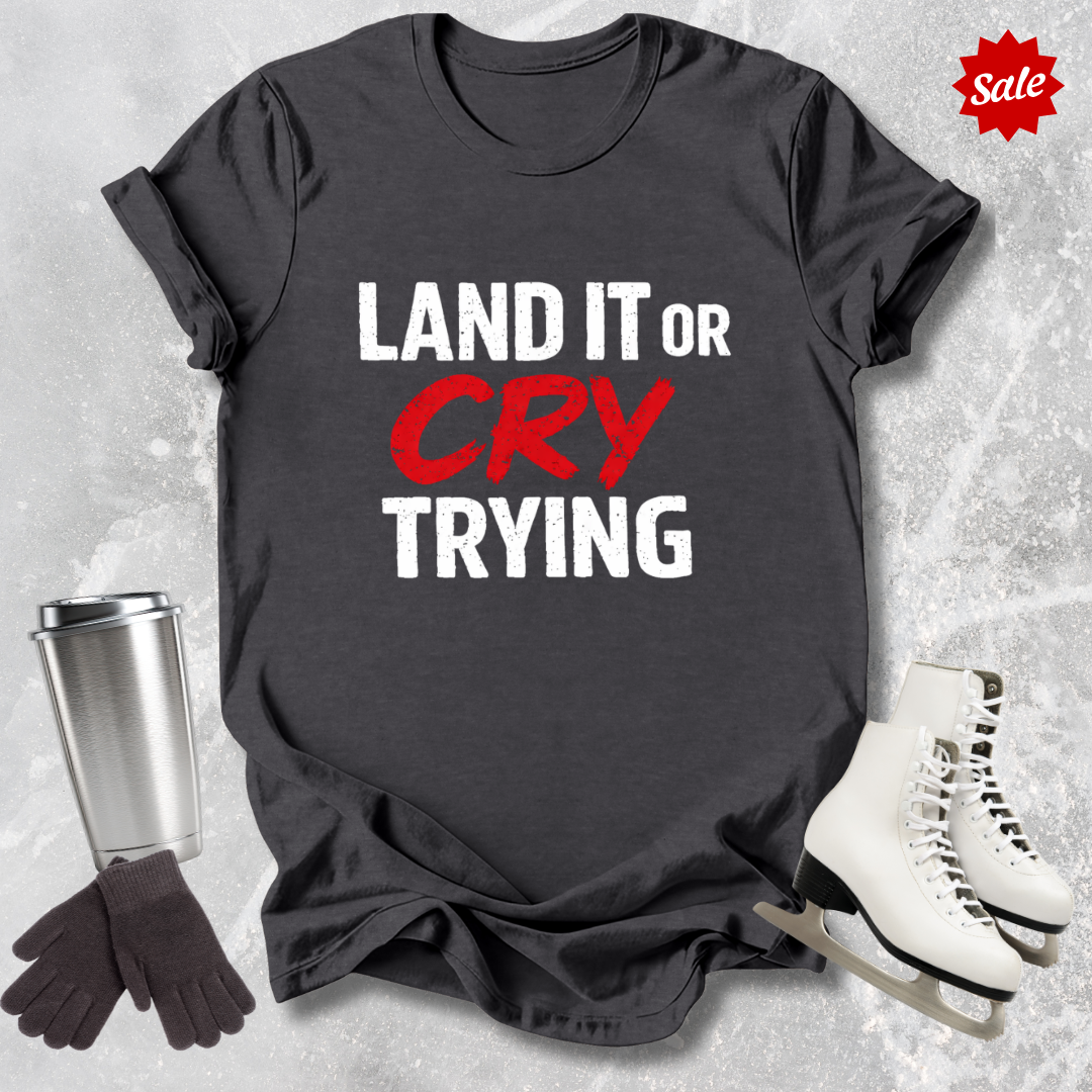 Gray t-shirt with 'LAND IT OR CRY TRYING' text, next to a silver tumbler, black gloves, and white skates on a textured gray background.