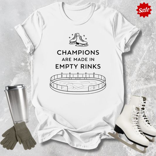 White t-shirt with a figure skating design and Champions are Made in Empty Rinks text, surrounded by ice skates and a tumbler on a textured surface.