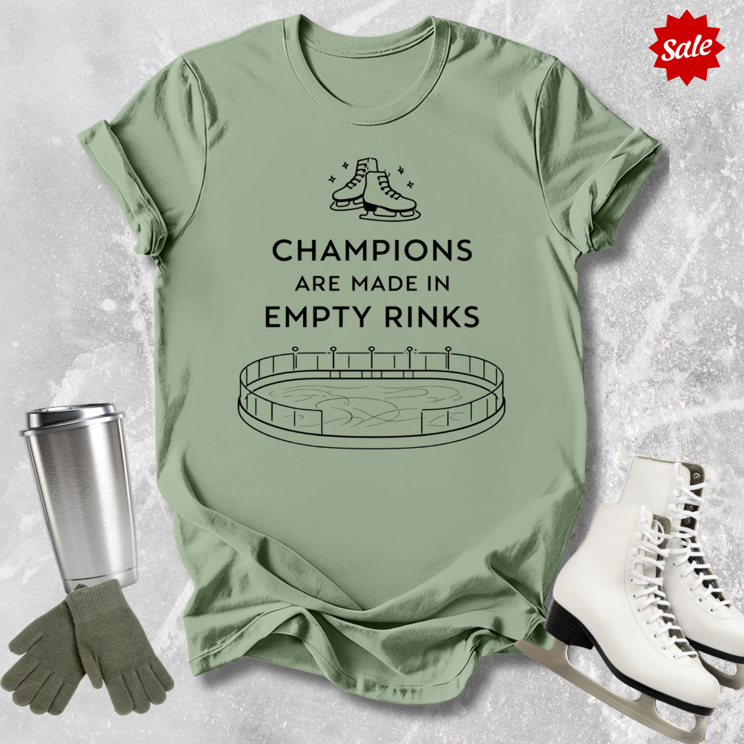 Green t-shirt with a figure skating design and Champions are Made in Empty Rinks text, surrounded by ice skates and a tumbler on a textured surface.
