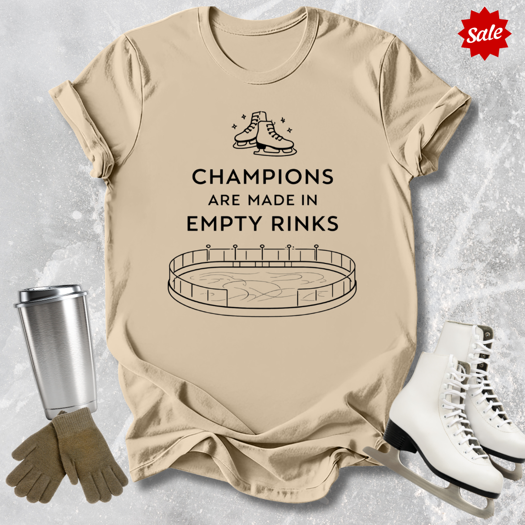 Beige t-shirt with a figure skating design and Champions are Made in Empty Rinks text, surrounded by ice skates and a tumbler on a textured surface.