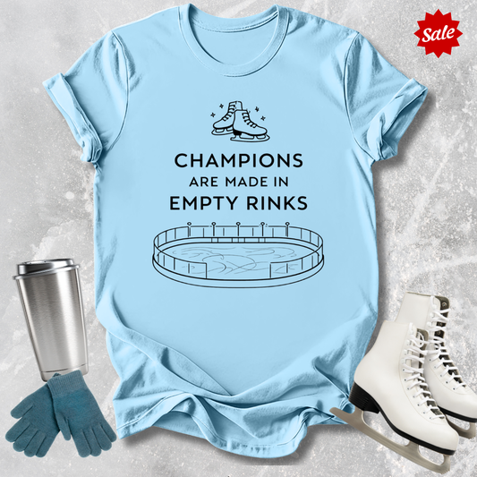 Light blue t-shirt with a figure skating design and Champions are Made in Empty Rinks text, surrounded by ice skates and a tumbler on a textured surface.