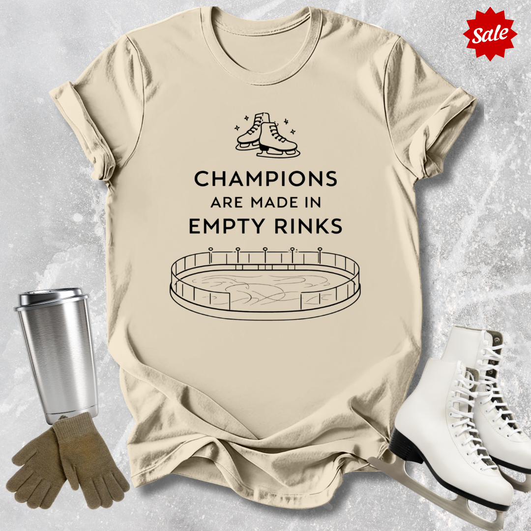 Beige t-shirt with a figure skating design and Champions are Made in Empty Rinks text, surrounded by ice skates and a tumbler on a textured surface.
