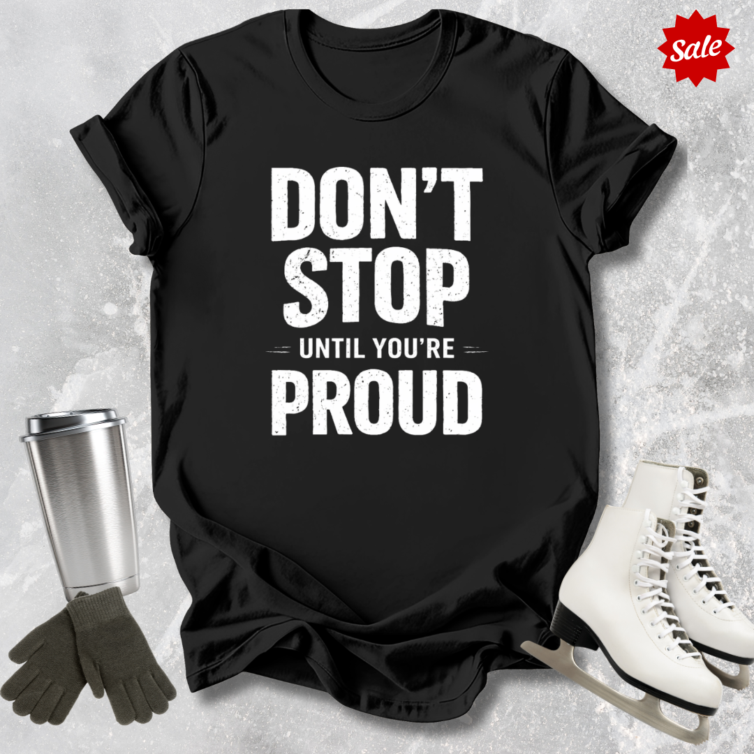Black t-shirt with 'Don't Stop Until You're Proud' text, surrounded by a silver tumbler, black gloves, and white skates on a textured gray background.