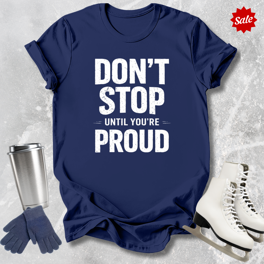 Navy blue t-shirt with 'Don't Stop Until You're Proud' text, next to a silver tumbler, gloves, and white skates on a textured gray background.