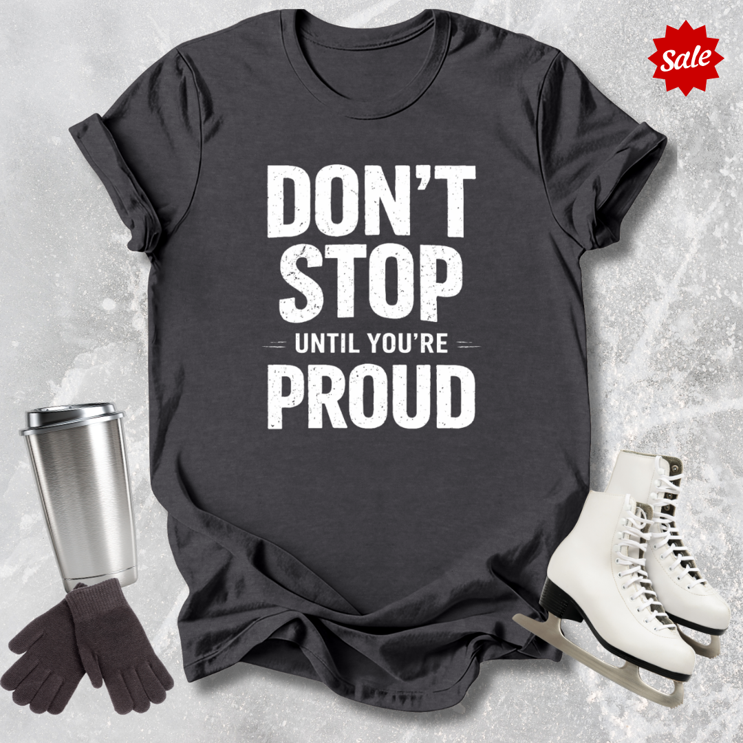 Black t-shirt with 'Don't Stop Until You're Proud' text, surrounded by a silver tumbler, black gloves, and white skates on a textured gray background.
