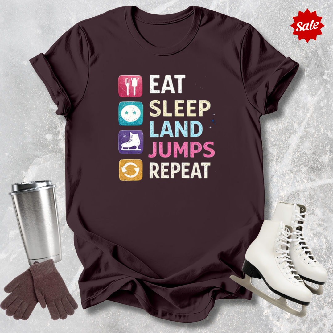 Brown t-shirt with 'Eat Sleep Land Jumps Repeat' text and graphics, next to a silver tumbler, gloves, and white skates on a textured gray background.
