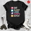 Black t-shirt with colorful graphic and text 'Eat Sleep Land Jumps Repeat' on a textured surface with ice skates and a tumbler.