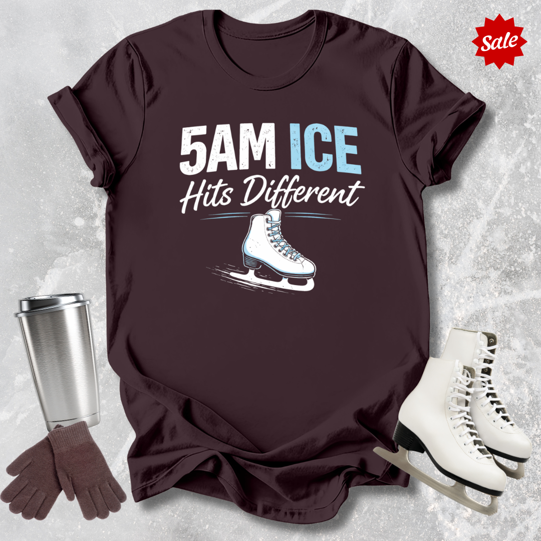Brown t-shirt with '5AM ICE Hits Different' text and ice skate graphic, surrounded by ice skates, gloves, and a tumbler on a textured gray background.