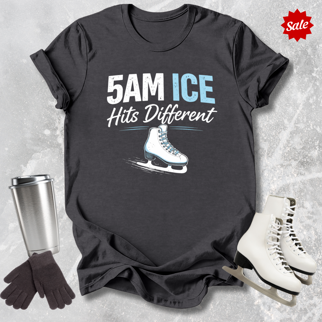 Black t-shirt with '5AM ICE Hits Different' text and ice skate graphic, surrounded by ice skates and a tumbler on a textured gray background.