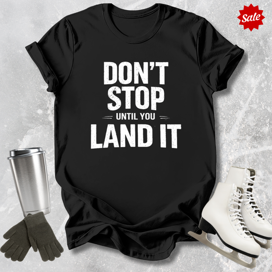 Black t-shirt with 'Don't Stop Until You Land It' text, next to a silver tumbler, black gloves, and white skates on a textured gray background.