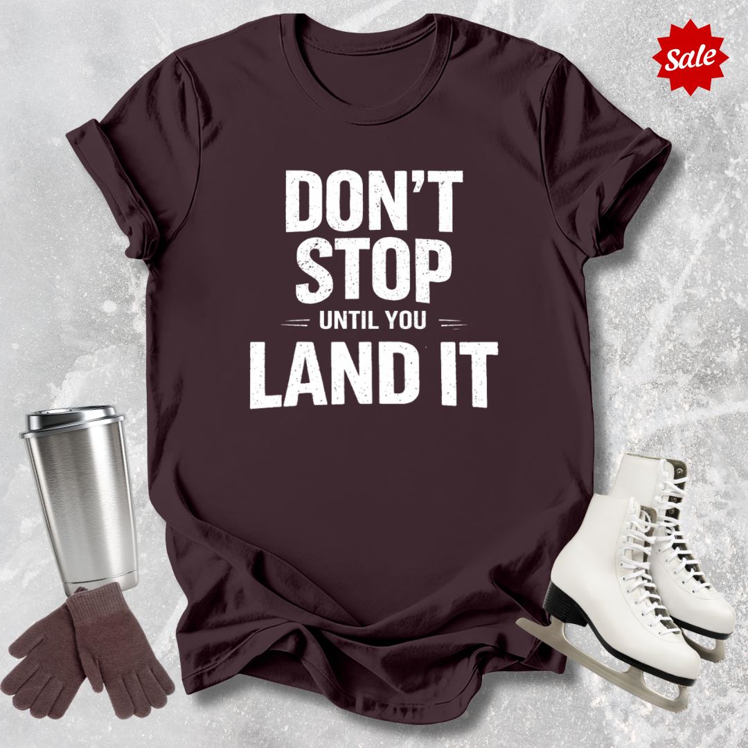 Maroon t-shirt with motivational quote, silver tumbler, brown gloves, and white skates on a textured gray background.