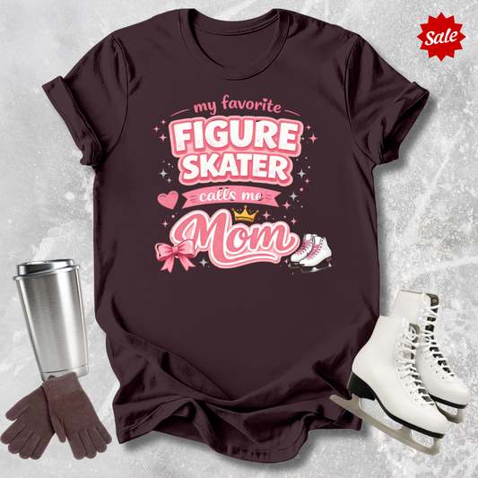 Maroon t-shirt with 'my favorite figure skater calls me mom' text, surrounded by ice skates, gloves, and a tumbler on a textured gray background.