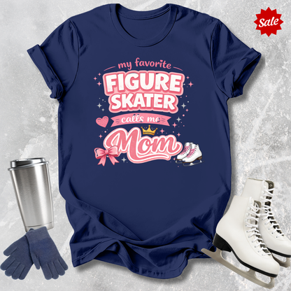 Navy blue t-shirt with pink text 'My Favorite Figure Skater Calls Me Mom' on a textured gray background with ice skates and a tumbler.