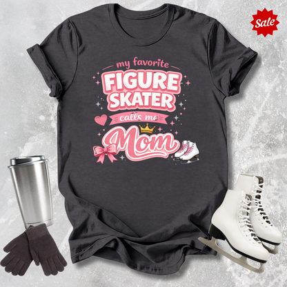 Black t-shirt with pink text 'My favorite figure skater calls me Mom' on a textured gray background.