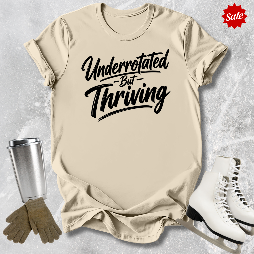 Beige t-shirt with 'Underrotated But Thriving' text, silver tumbler, brown gloves, and white sneakers on a textured gray background.