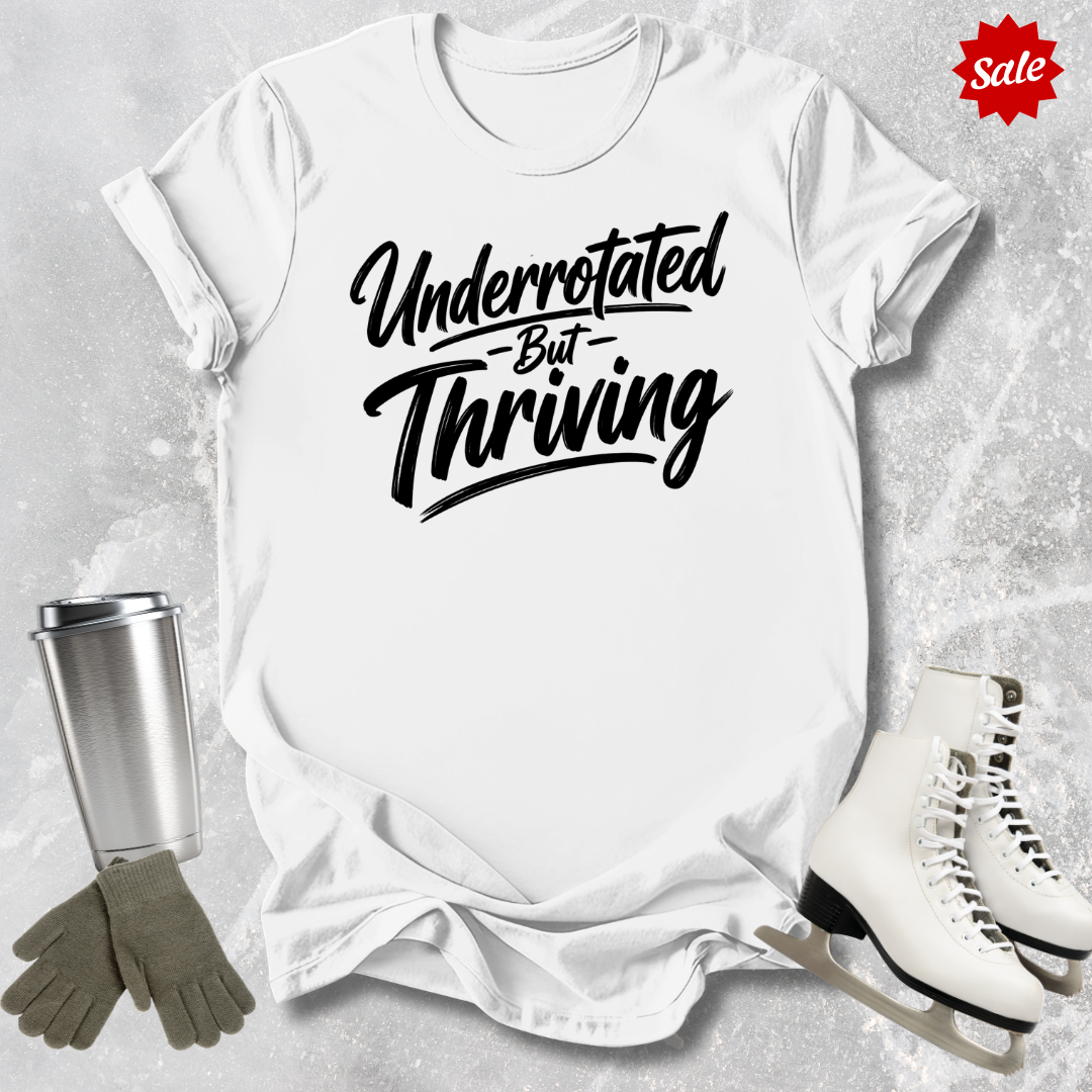 White t-shirt with 'Underrotated But Thriving' text, surrounded by a silver tumbler, gray gloves, and white skates on a textured surface.