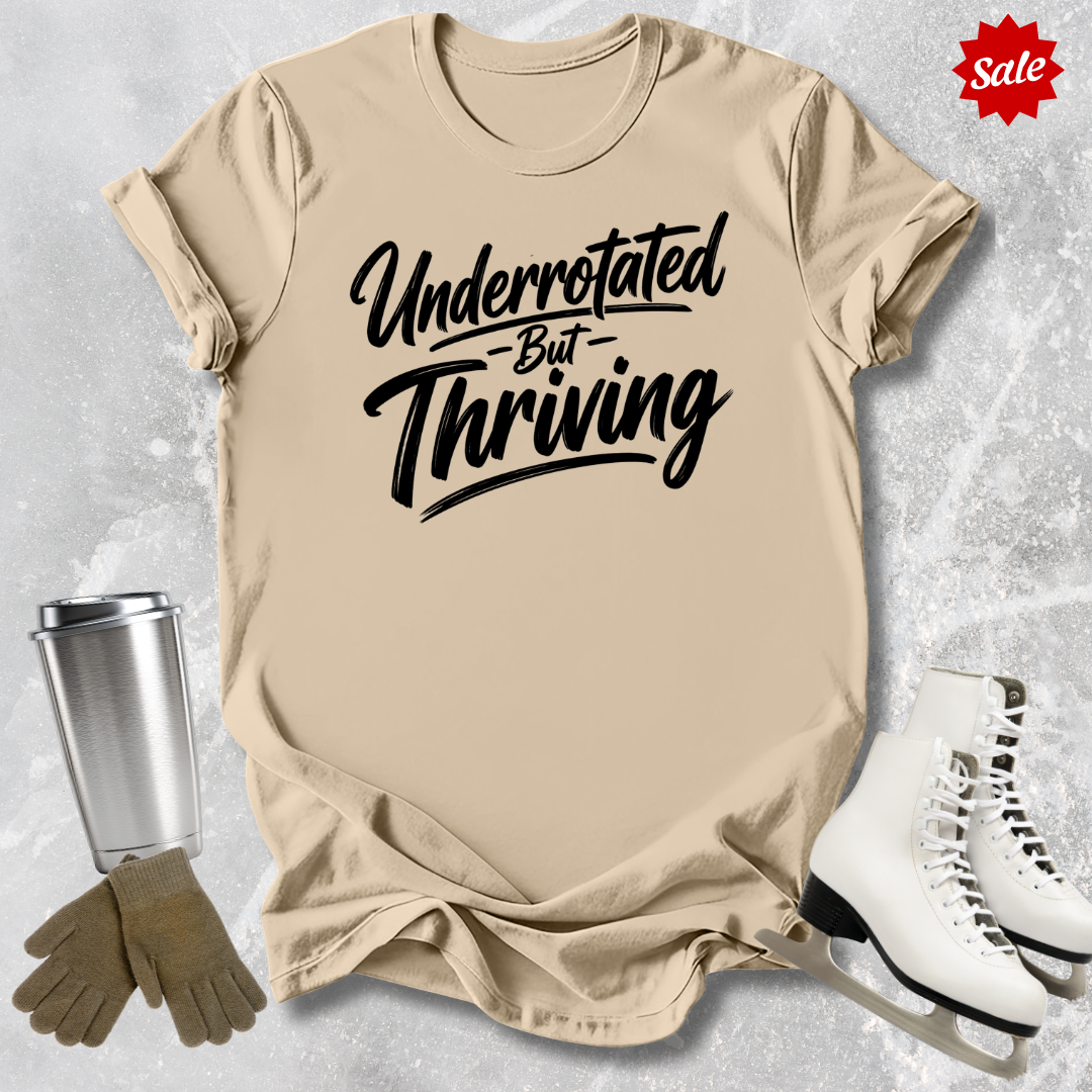 Beige t-shirt with 'Underrotated But Thriving' text, silver tumbler, brown gloves, and white skates on a light gray background.