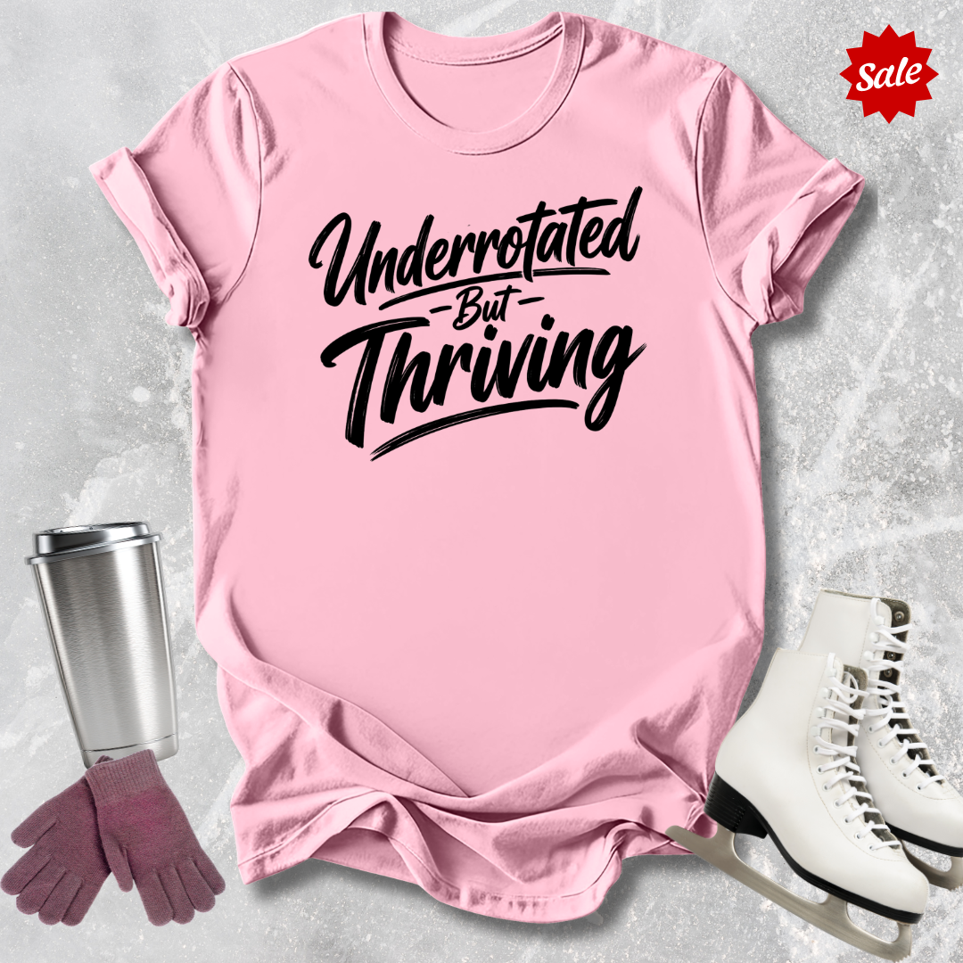 Pink t-shirt with 'Underrotated But Thriving' text, surrounded by a silver tumbler, pink gloves, and white skates on a light gray background.