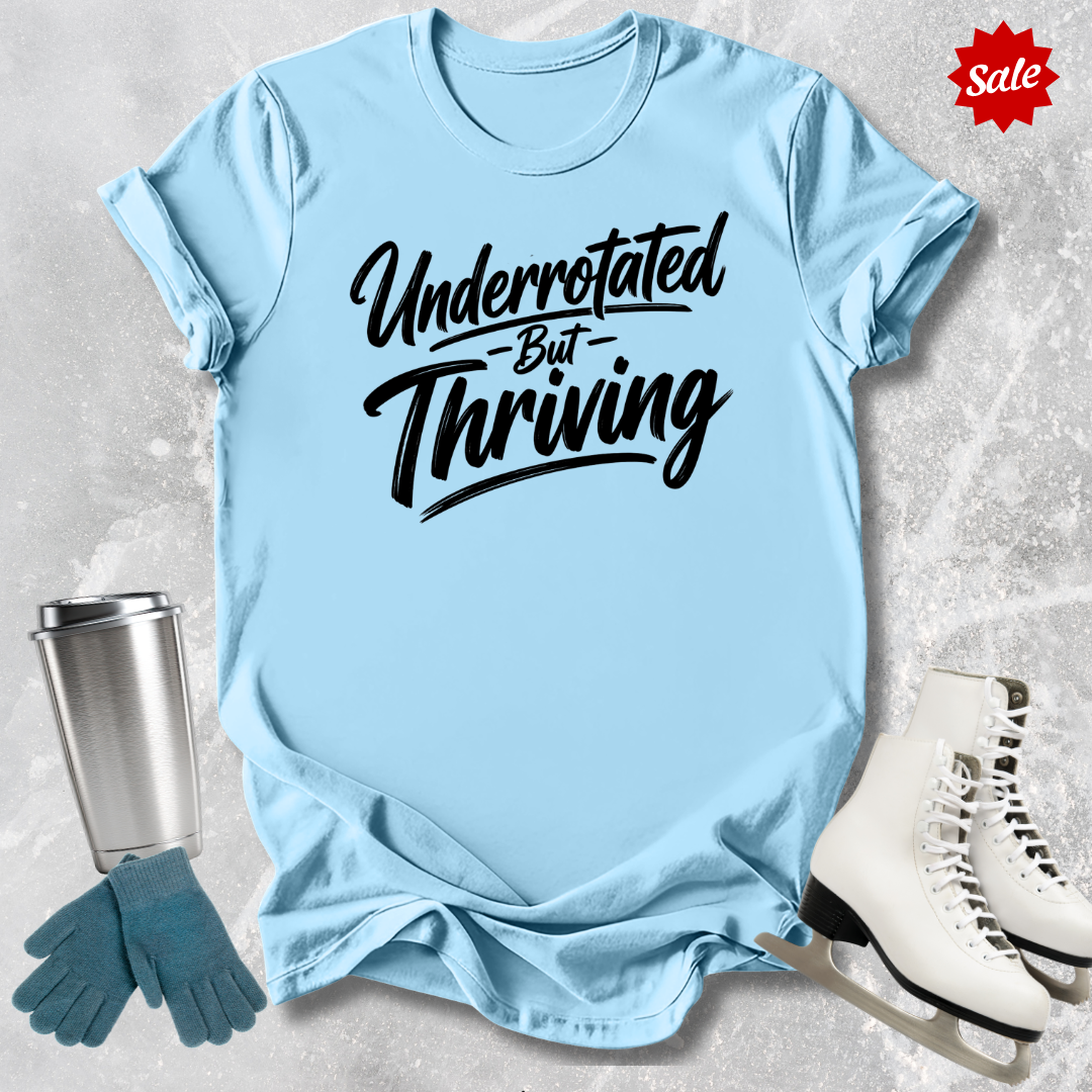 Light blue t-shirt with 'Underrotated But Thriving' text, next to a silver tumbler, green gloves, and white skates on a textured surface.