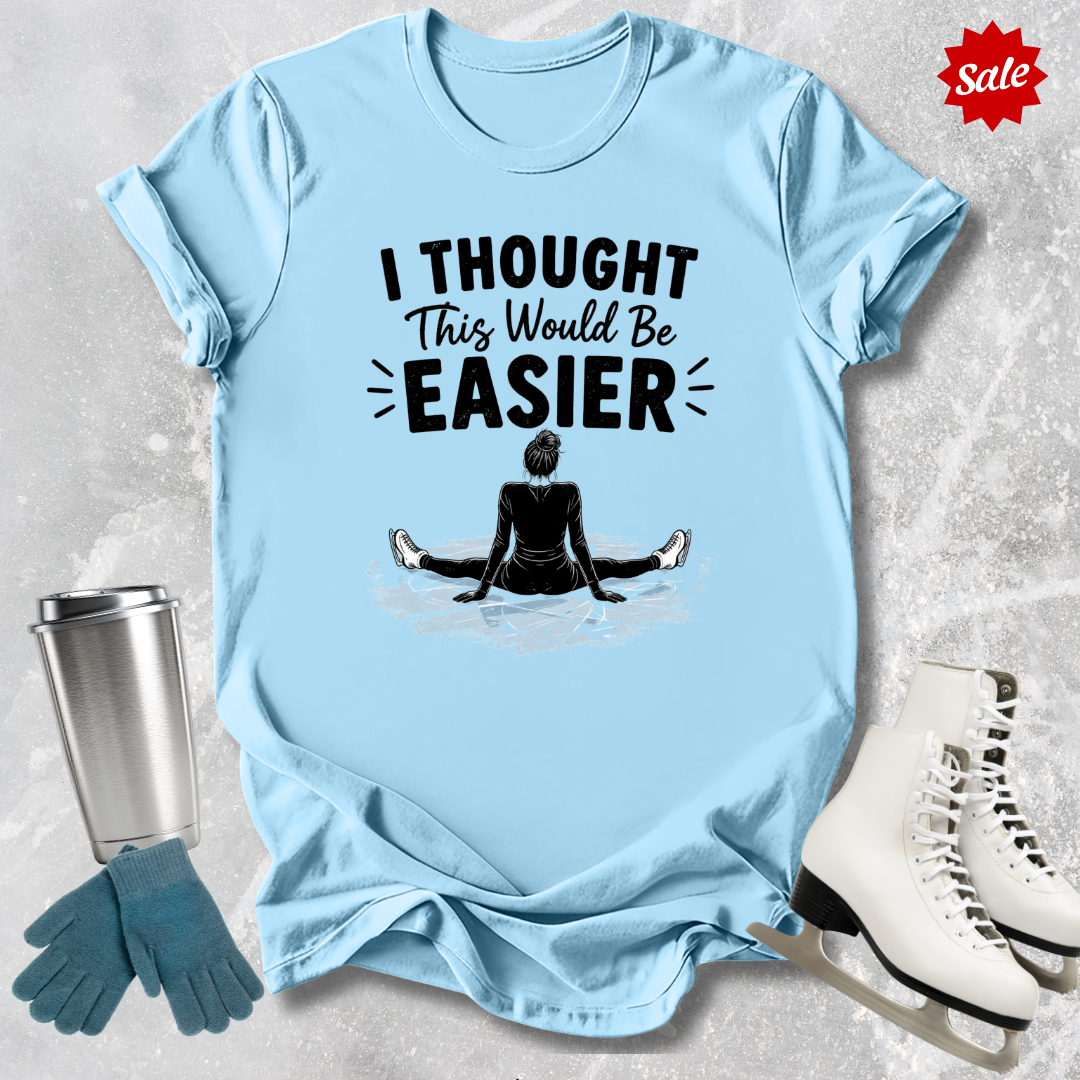 Light blue t-shirt with 'I Thought This Would Be Easier' text and graphic, surrounded by a metal tumbler, blue gloves, and white ice skates on a textured surface.