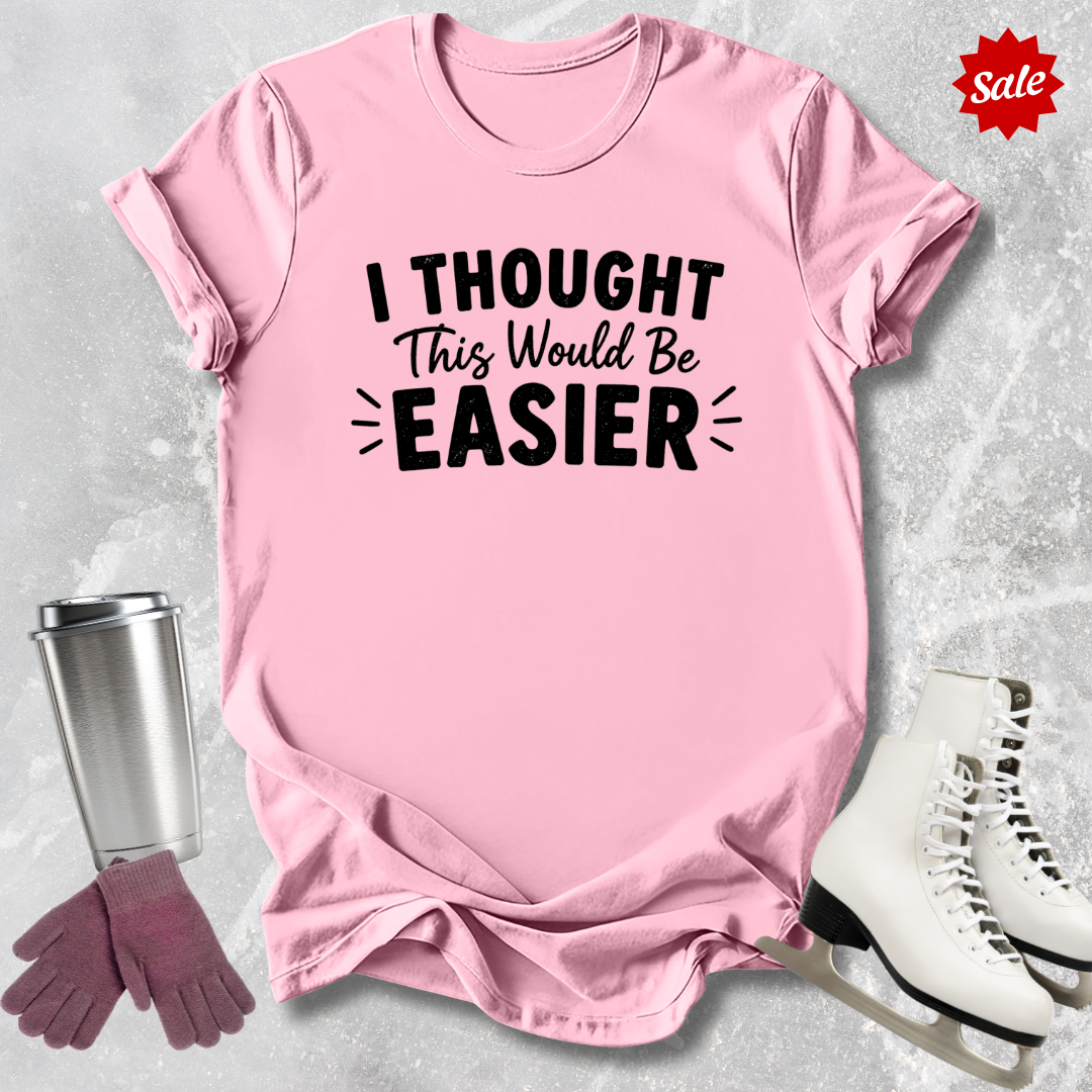 Pink t-shirt with 'I Thought This Would Be Easier' text, surrounded by a silver tumbler, pink gloves, and white skates on a light gray background.