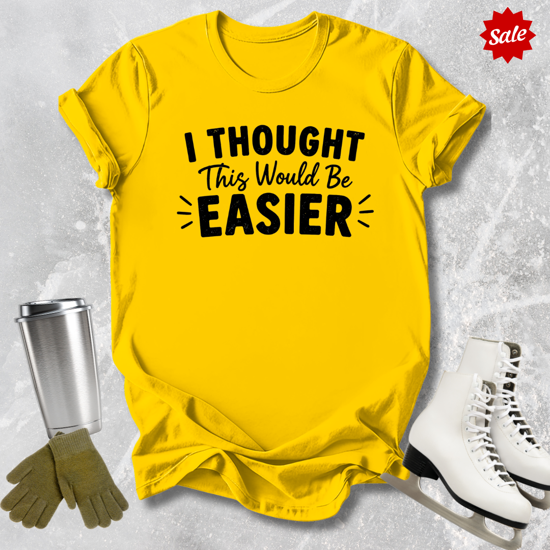 Yellow t-shirt with 'I Thought This Would Be Easier' text, surrounded by a tumbler, gloves, and skates on a textured gray background.