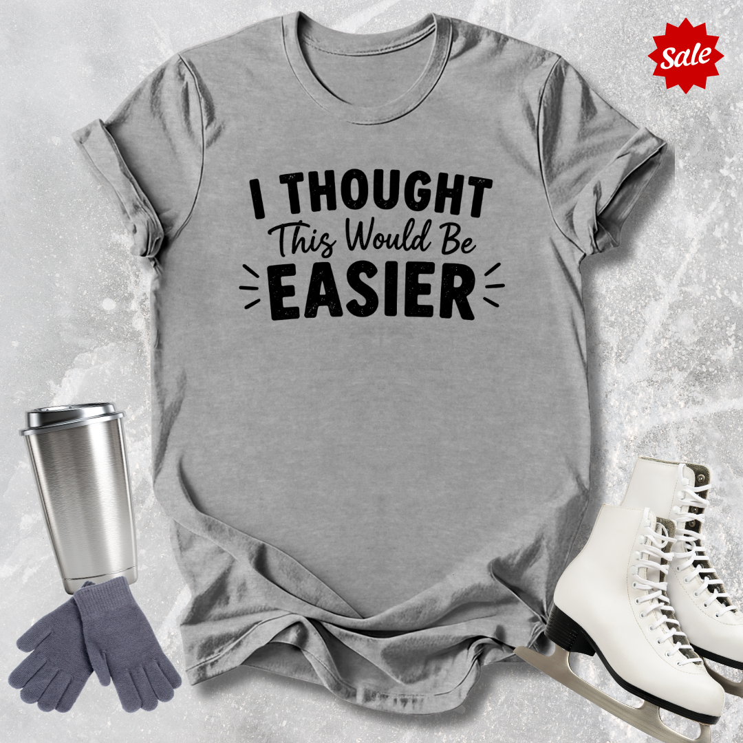 Gray t-shirt with 'I Thought This Would Be Easier' text, surrounded by a silver tumbler, gray gloves, and white skates on a textured surface.