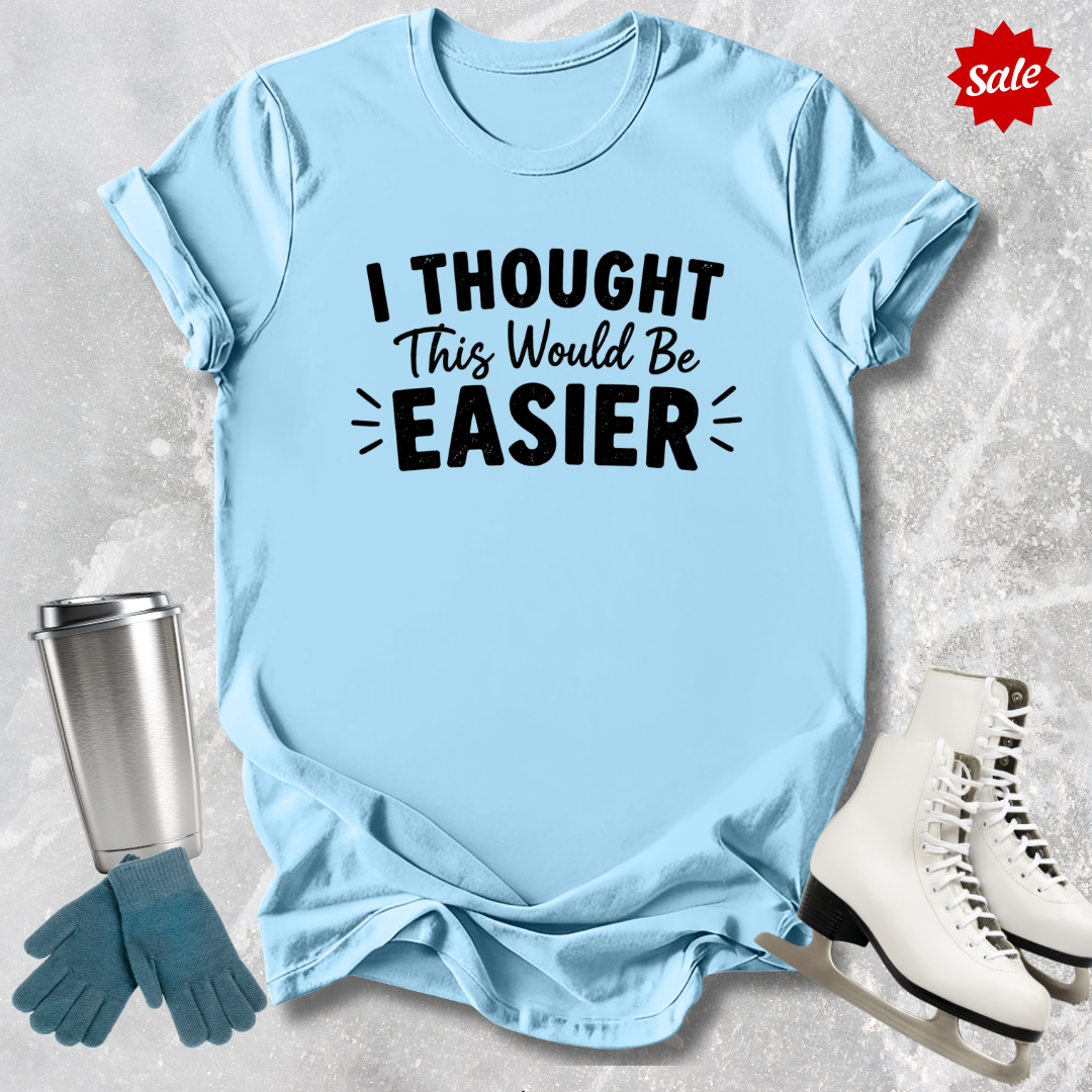 Light blue t-shirt with 'I Thought This Would Be Easier' text, next to a silver tumbler, green gloves, and white skates on a textured surface.