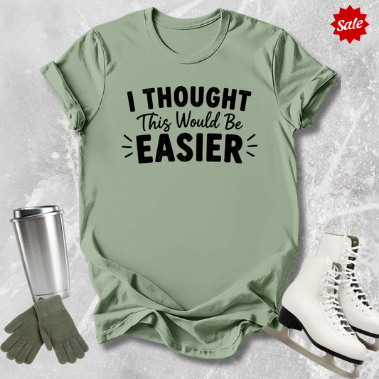 Green t-shirt with 'I Thought This Would Be Easier' text, next to a silver tumbler, green gloves, and white skates on a textured gray background.
