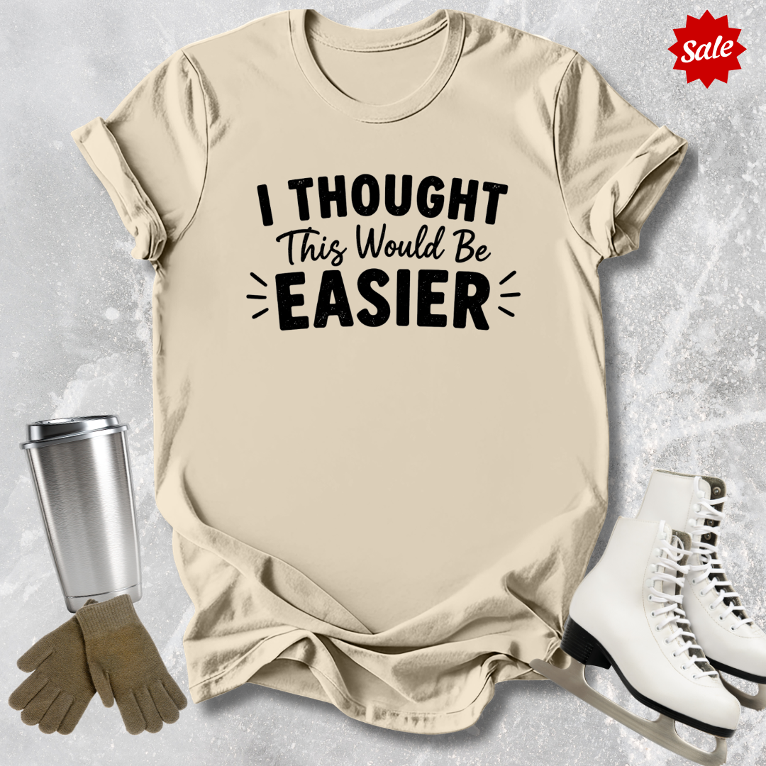 Beige t-shirt with 'I Thought This Would Be Easier' text, next to a silver tumbler, brown gloves, and white skates on a light gray background.