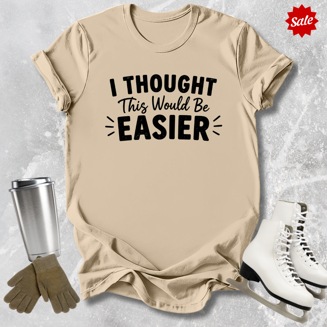 Beige t-shirt with 'I Thought This Would Be Easier' text, next to a silver tumbler, brown gloves, and white skates on a light gray background.