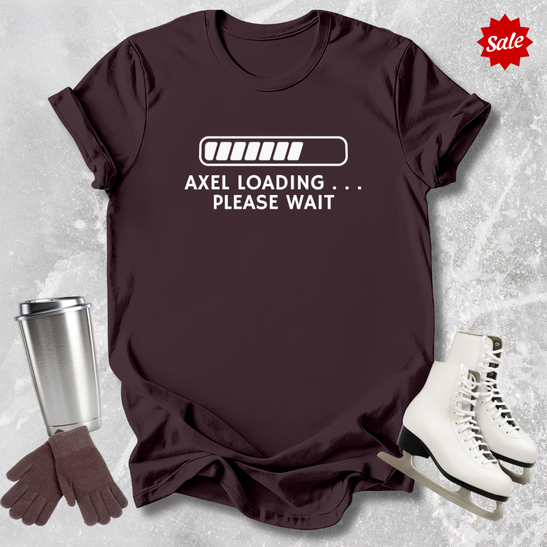 Brown t-shirt with 'Axel Loading Please Wait' text, next to a silver tumbler, gloves, and white skates on a textured gray background.