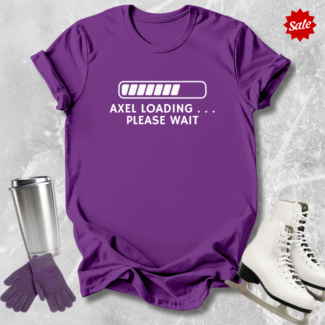Purple t-shirt with 'Axel Loading Please Wait' text, next to a silver tumbler, gloves, and white skates on a textured gray background.