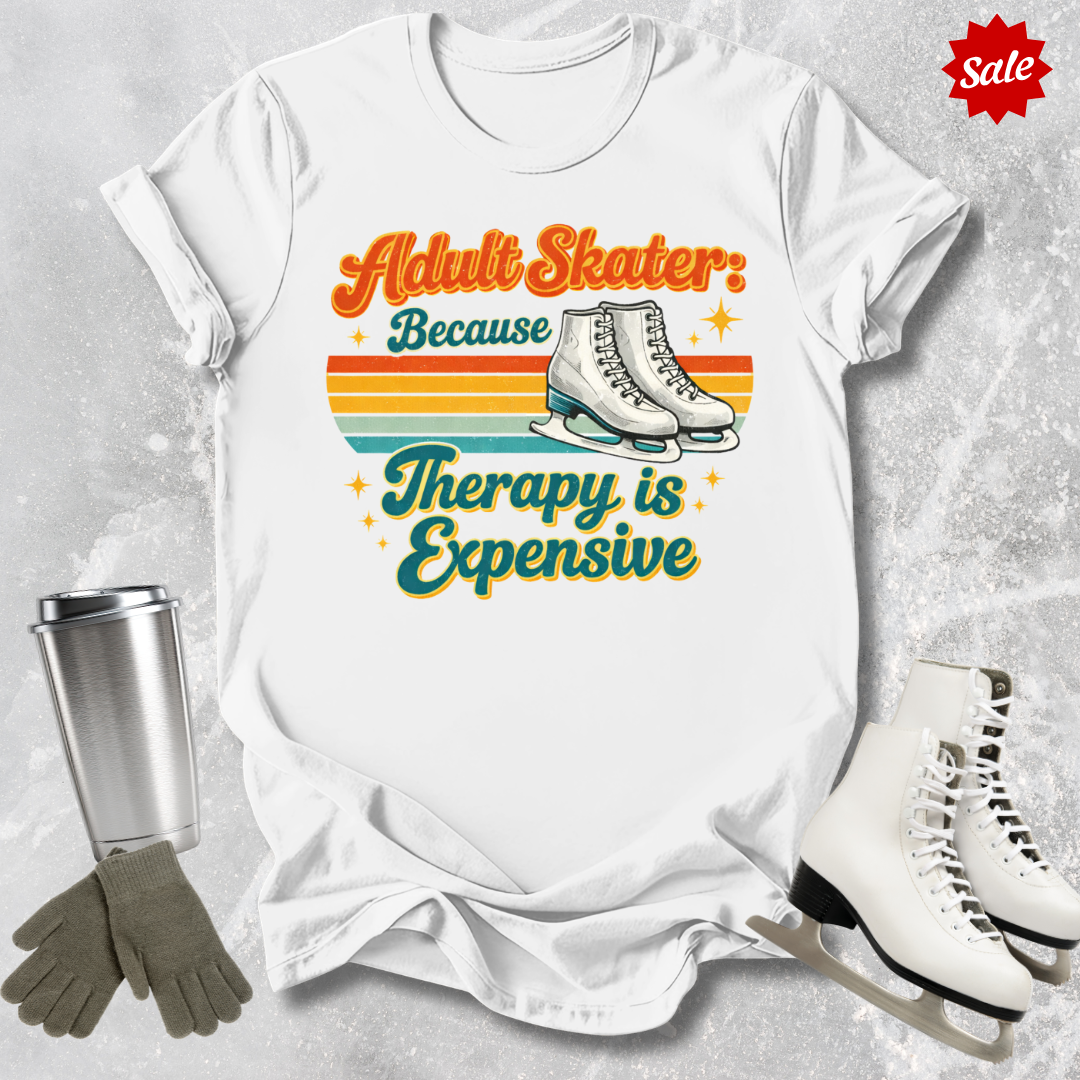 White t-shirt with 'Adult Skater Because Therapy is Expensive' graphic, surrounded by ice skates and a tumbler on a textured surface.