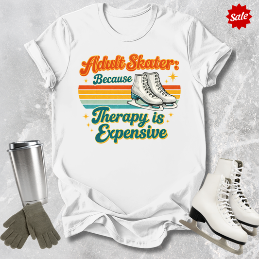 White t-shirt with 'Adult Skater Because Therapy is Expensive' graphic, surrounded by ice skates and a tumbler on a textured surface.