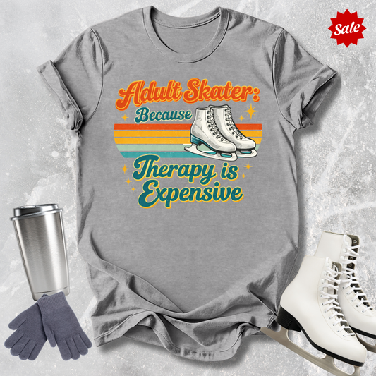 Gray t-shirt with 'Adult Skater Because Therapy is Expensive' text, surrounded by ice skates, gloves, and a tumbler on a textured gray background.