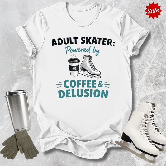 White t-shirt with 'Adult Skater: Powered by Coffee & Delusion' text, surrounded by ice skates and a tumbler on a textured surface.