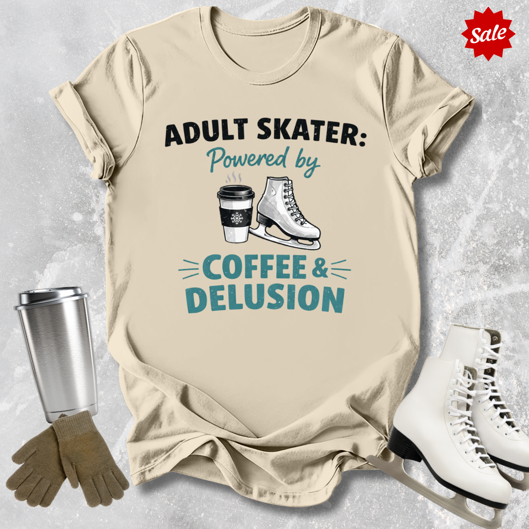 Beige t-shirt with text 'Adult Skater: Powered by Coffee & Delusion' on a textured surface with ice skates and a tumbler.