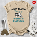 Beige t-shirt with 'Adult Skater: Powered by Coffee & Delusion' text, next to ice skates and a tumbler on a textured surface.