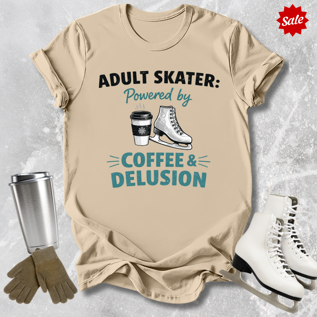 Beige t-shirt with 'Adult Skater: Powered by Coffee & Delusion' text, next to ice skates and a tumbler on a textured surface.