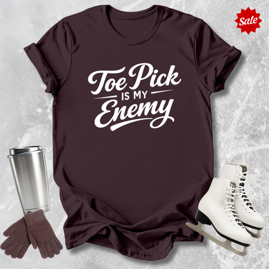Brown t-shirt with 'Toe Pick is My Enemy' text, silver tumbler, brown gloves, and white skates on a textured gray background.