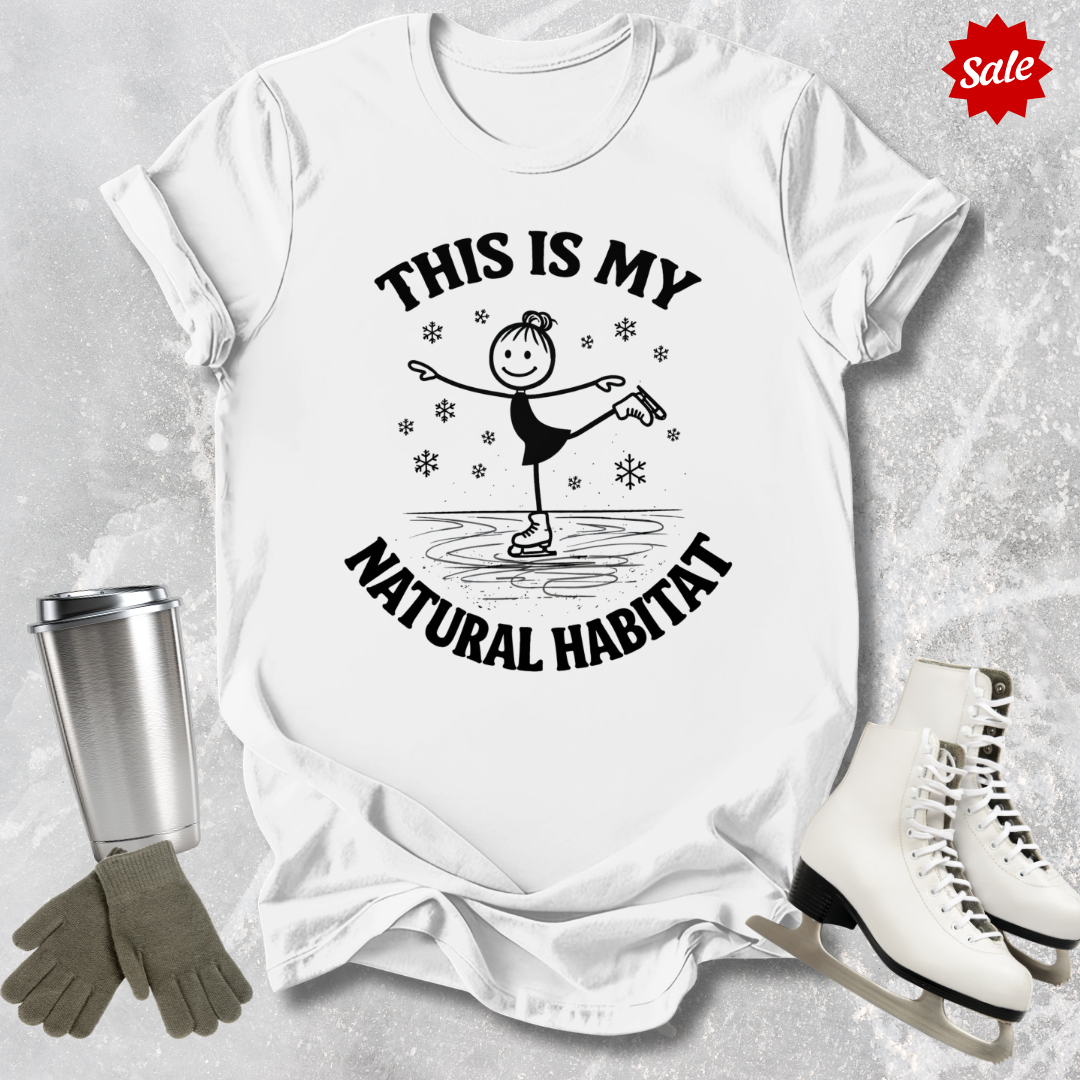 White t-shirt with 'This is my natural habitat' graphic, surrounded by ice skates, gloves, and a tumbler on a textured surface.