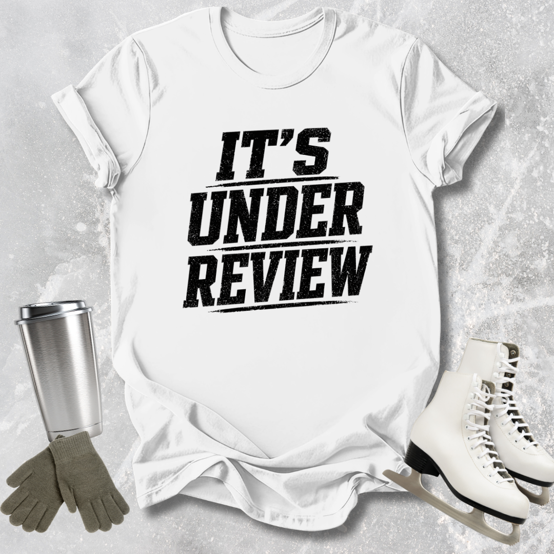 White t-shirt with 'IT'S UNDER REVIEW' text, silver tumbler, gray gloves, and white skates on a light gray background