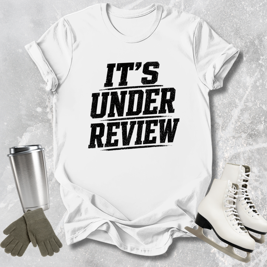 White t-shirt with 'IT'S UNDER REVIEW' text, silver tumbler, gray gloves, and white skates on a light gray background
