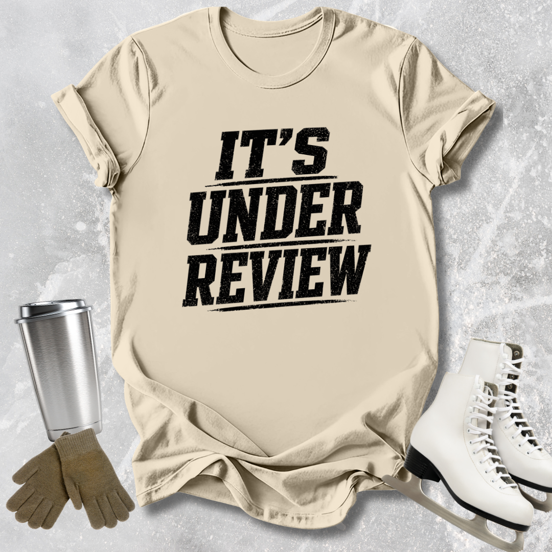 Beige t-shirt with 'IT'S UNDER REVIEW' text, next to a silver tumbler, brown gloves, and white shoes on a light gray background.