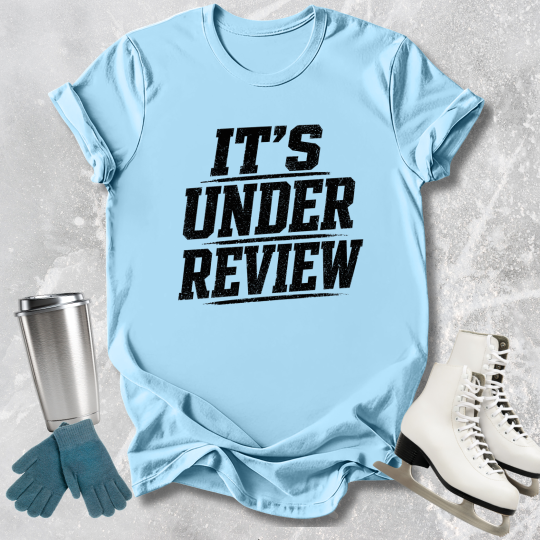 Light blue t-shirt with 'IT'S UNDER REVIEW' text, next to a silver tumbler, blue gloves, and white skates on a textured surface.