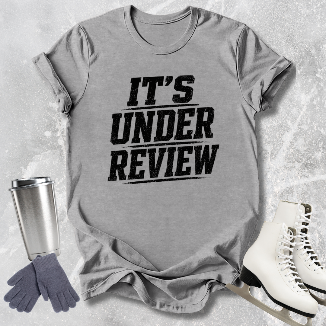 Gray t-shirt with 'IT'S UNDER REVIEW' text, next to a silver tumbler, gray gloves, and white skates on a light gray textured background.