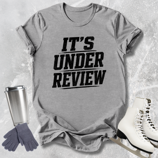 Gray t-shirt with 'IT'S UNDER REVIEW' text, next to a silver tumbler, gray gloves, and white skates on a light gray textured background.