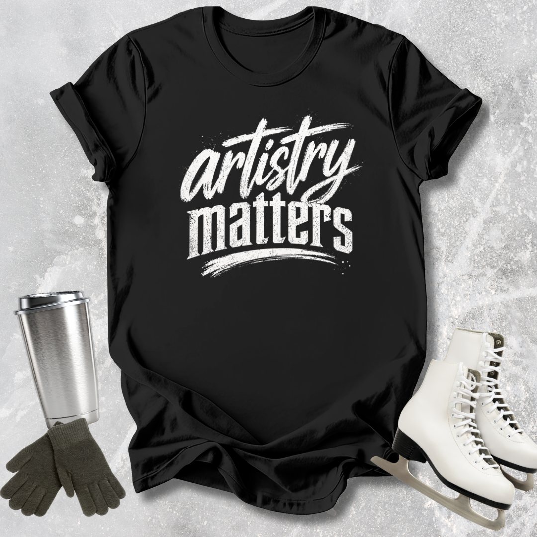 Black t-shirt with 'artistry matters' text, next to ice skates and a tumbler on a textured surface.