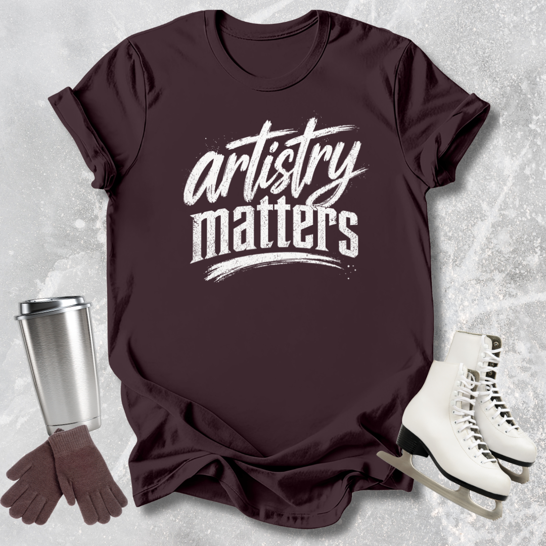 Maroon t-shirt with 'artistry matters' text, silver tumbler, brown gloves, and white skates on a textured gray background